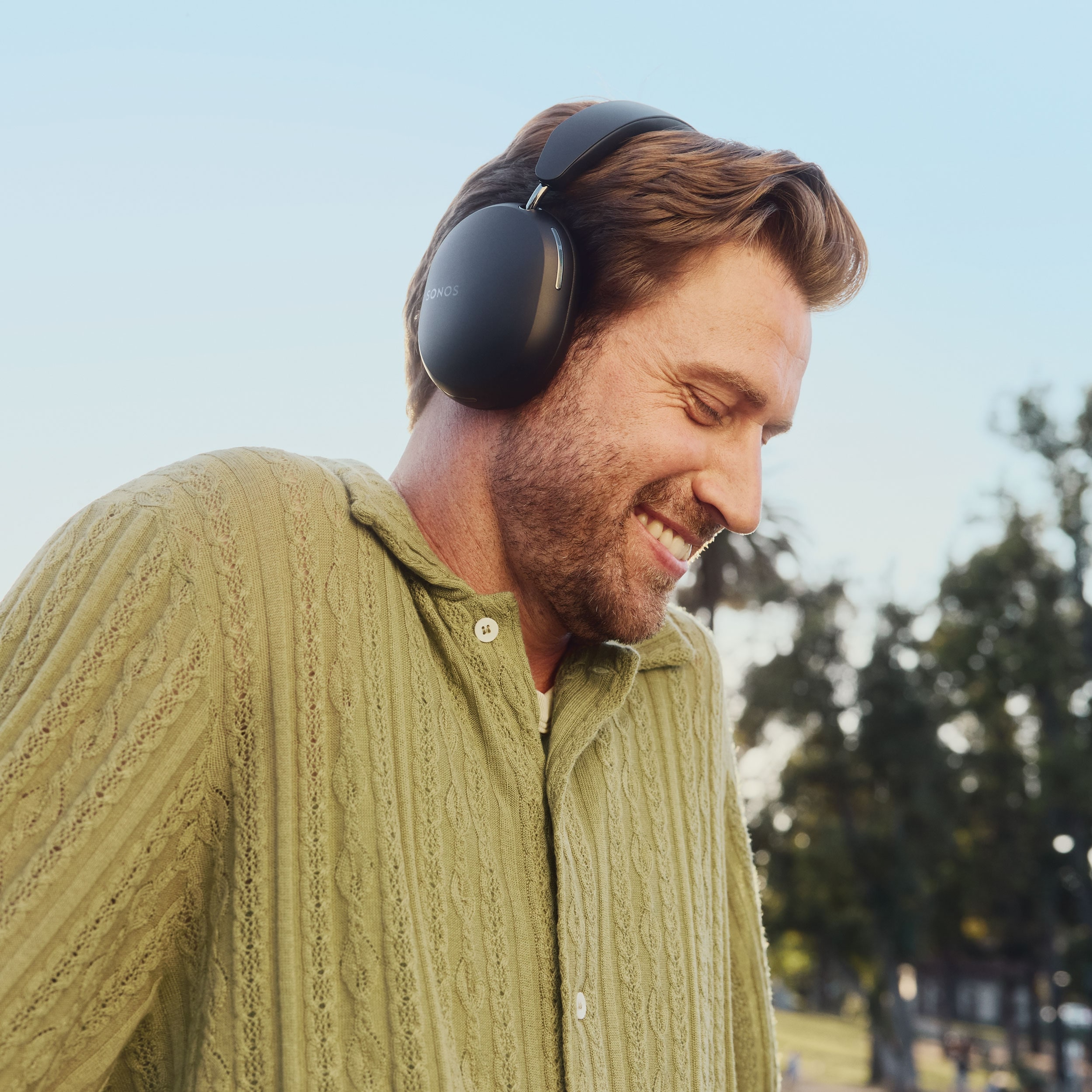 Una persona sorride mentre cammina nel parco ascoltando la musica con un paio di cuffie Sonos Ace nere