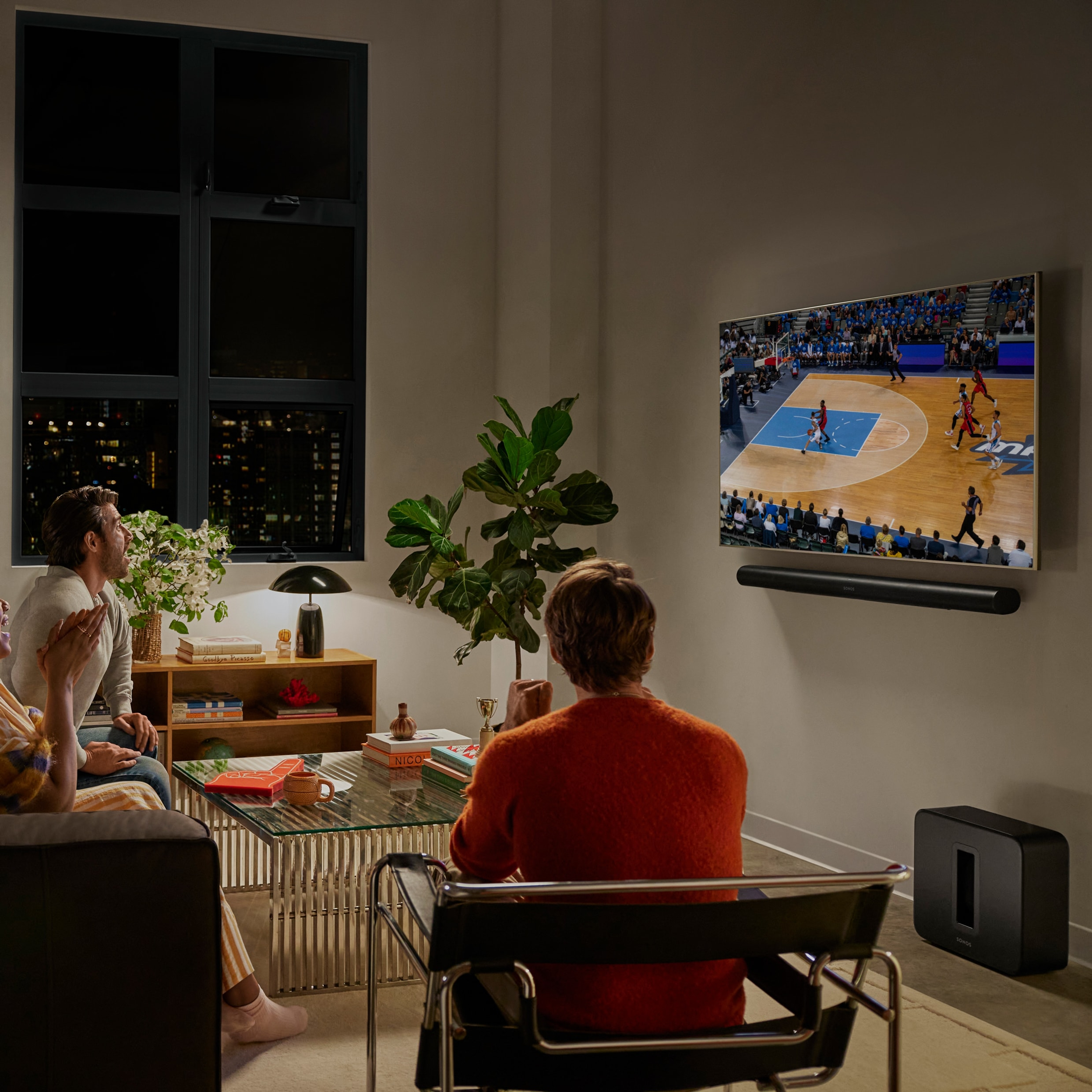En gruppe venner i en stue ser på en basketballkamp med en svart Arc Ultra og en svart Sub 4